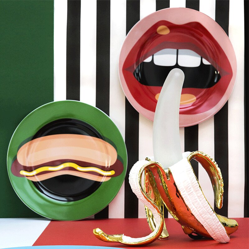 Two novelty plates with artistic designs of a hot dog and a banana, against a black and white striped background.