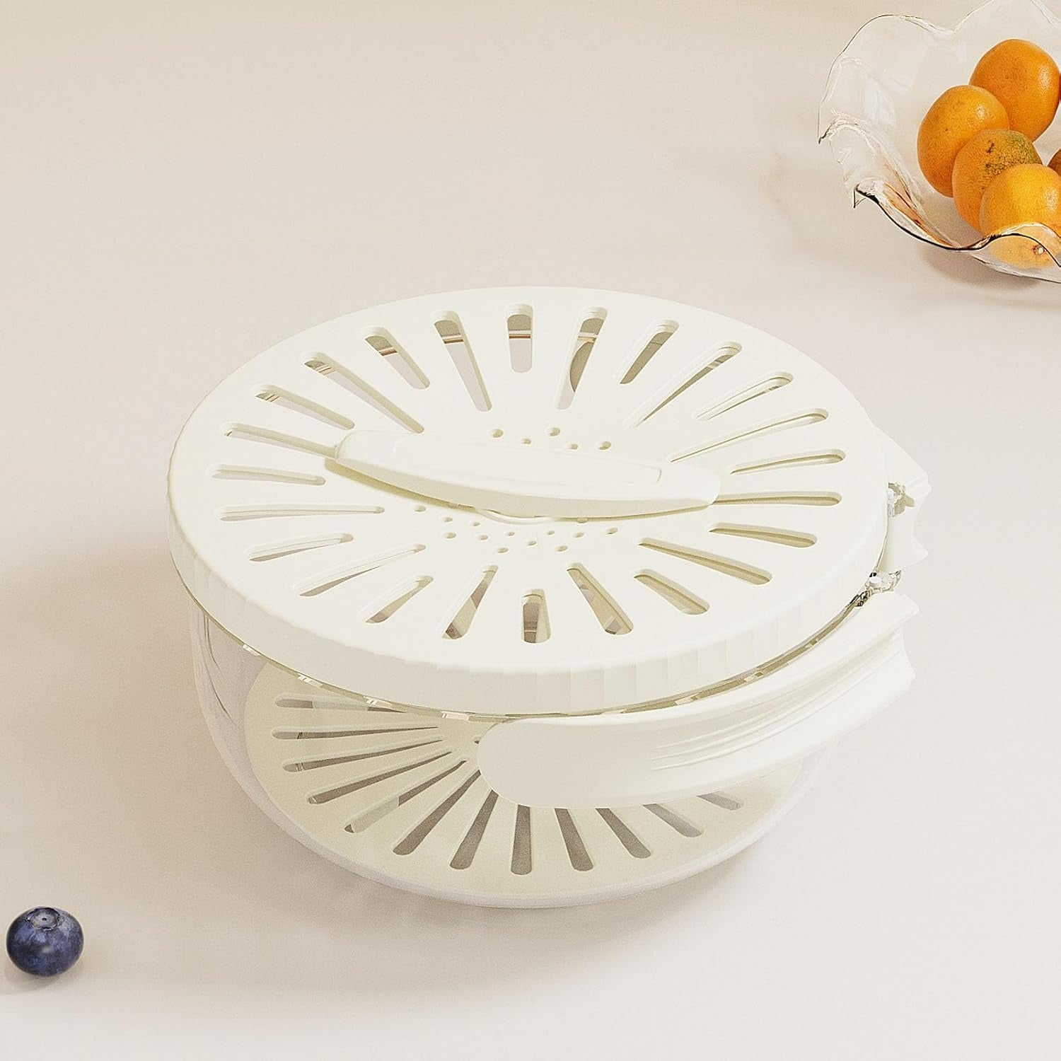 White kitchen steamer basket on a light beige surface with a small blueberry and a bowl of oranges in the background.