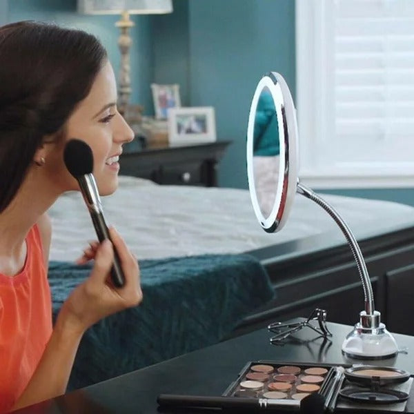 Woman applying makeup with a brush in front of a round, illuminated mirror on a desk.