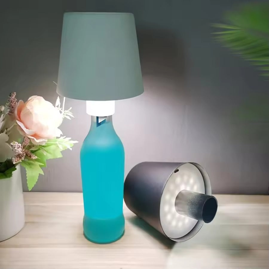 Blue bottle lamp with gray shade on a wooden surface next to a vase with flowers.