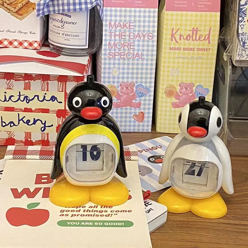 Two penguin-shaped calendars on a surface with various items in the background.