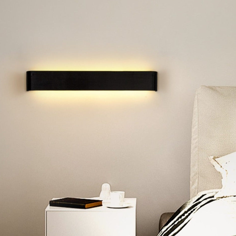 Modern wall light fixture on a neutral wall above a nightstand with a book and small round object.