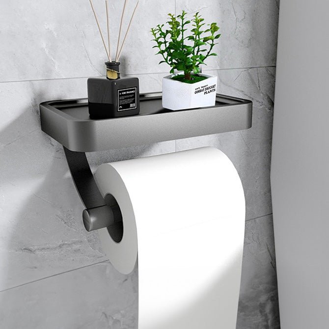 Wall-mounted toilet paper holder with a plant and decorative item on a gray tiled wall.